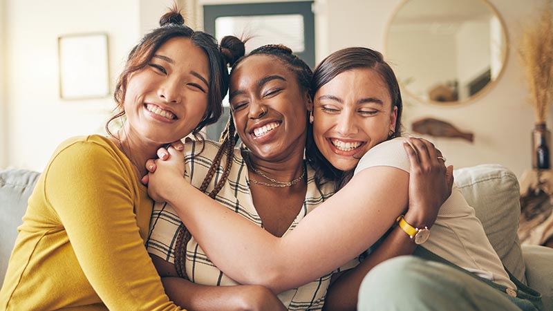 Portrait of a group of women, friends hug on sofa with smile and bonding in living room together in embrace. Hug, love and friendship, girls on couch with diversity, pride and people in home with fun Dentist in Kernersville, NC